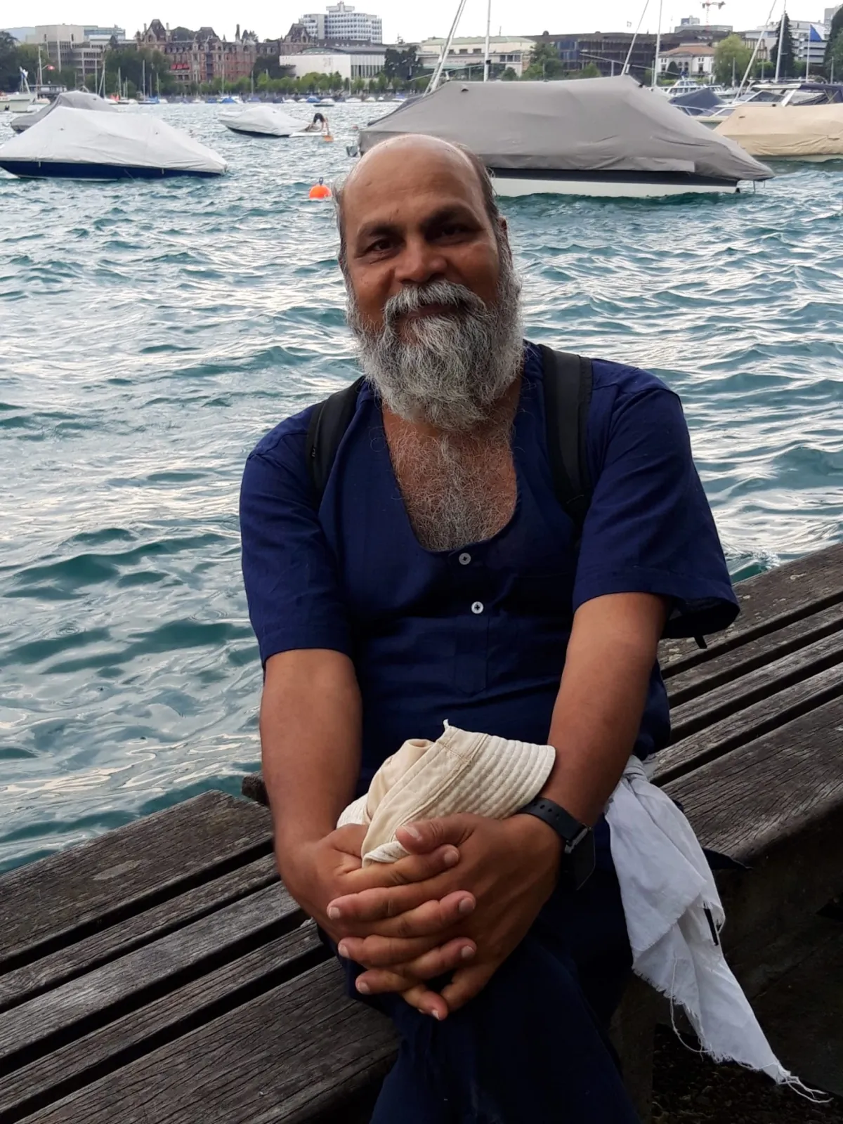 Swami-ji seated on a bench at a harbour, boats moored behind