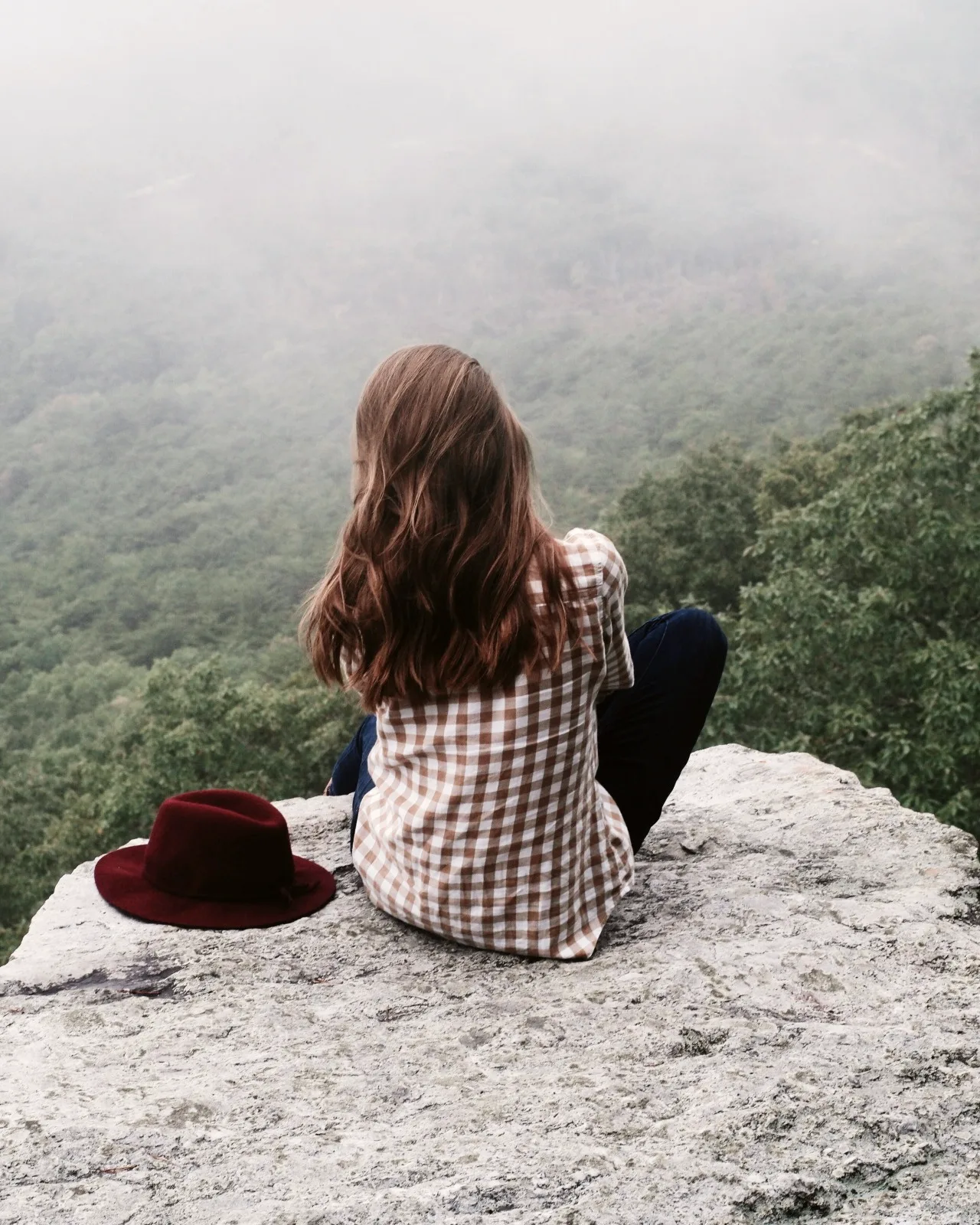 A woman sitting alone on a rock above a mist-filled forest valley