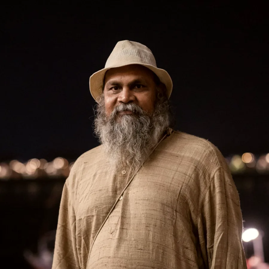 Swami Ashok Samdarshi standing on the ghats at night, the lights of the far bank behind him